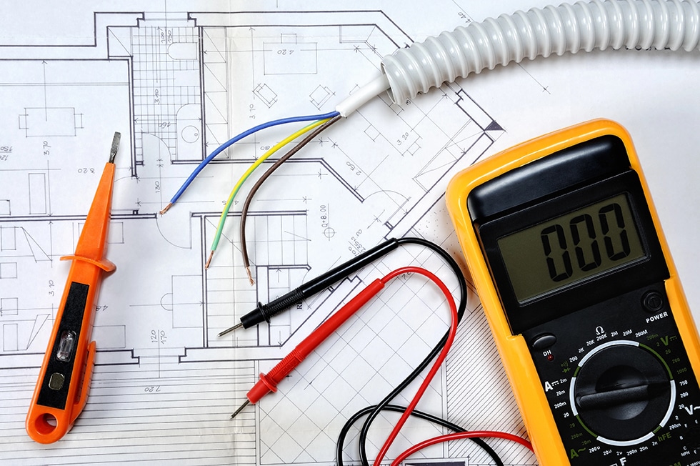 Top view of components and electrical equipment placed on the ho A digital multimeter, test leads, screwdriver, and exposed wires are placed on top of architectural blueprints for a building’s mechanical and electrical plan, reflecting the detailed preparation required for industrial and commercial projects.