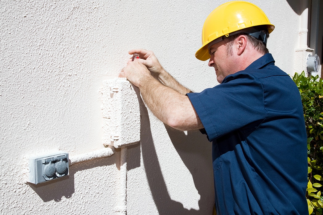 Electrical or Cable Repairman A man wearing a yellow hard hat and blue shirt uses a tool to work on an outdoor electrical box mounted on a textured wall in a residential or commercial area, with some greenery visible nearby.